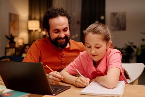 Smiling father guiding daughter during her homework session while sitting at table with laptop and books e engaging in educational activities in relaxed home environment